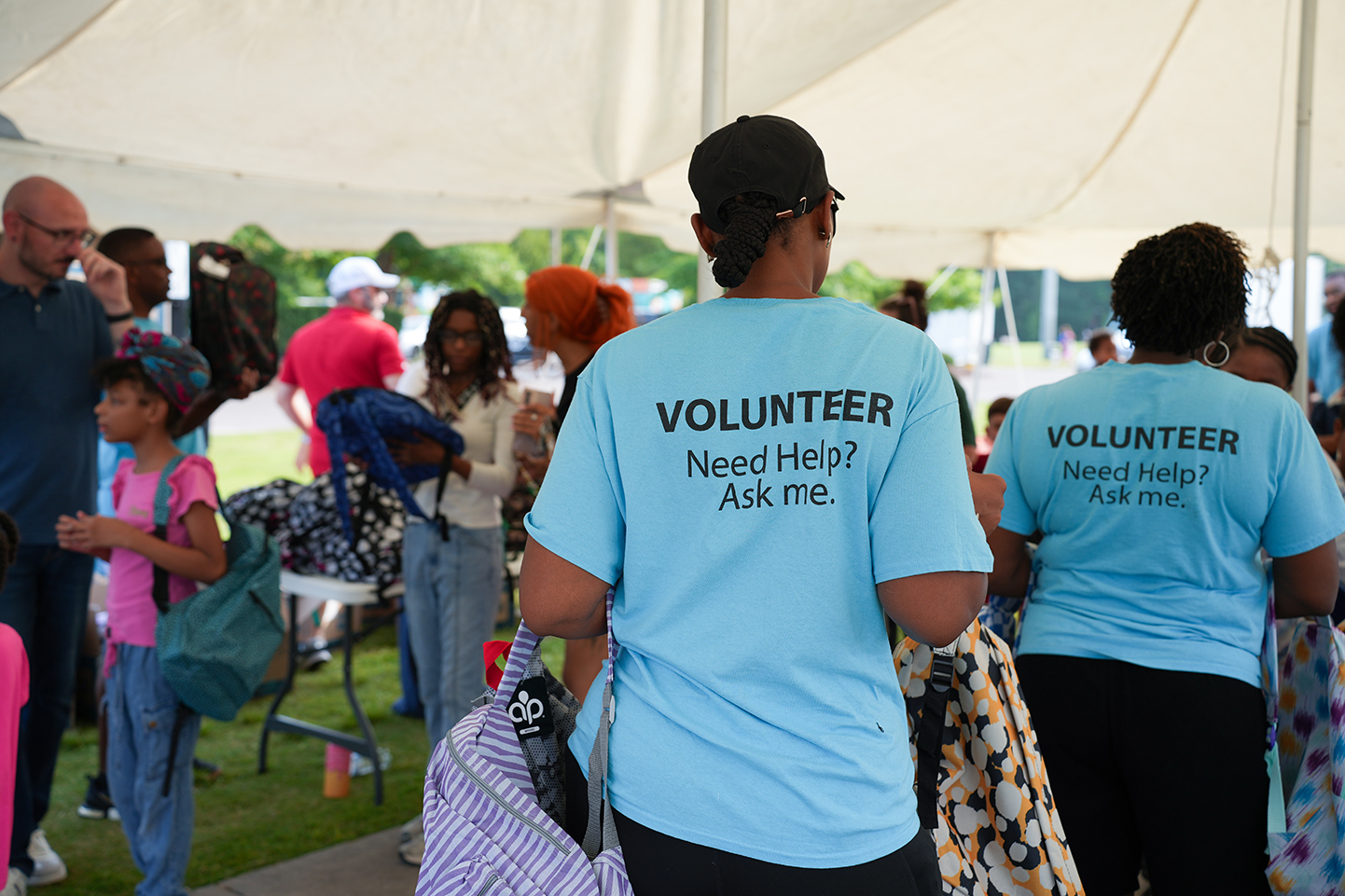 A group of volunteers in light blue shirts stands under a tent. One volunteer has a backpack.