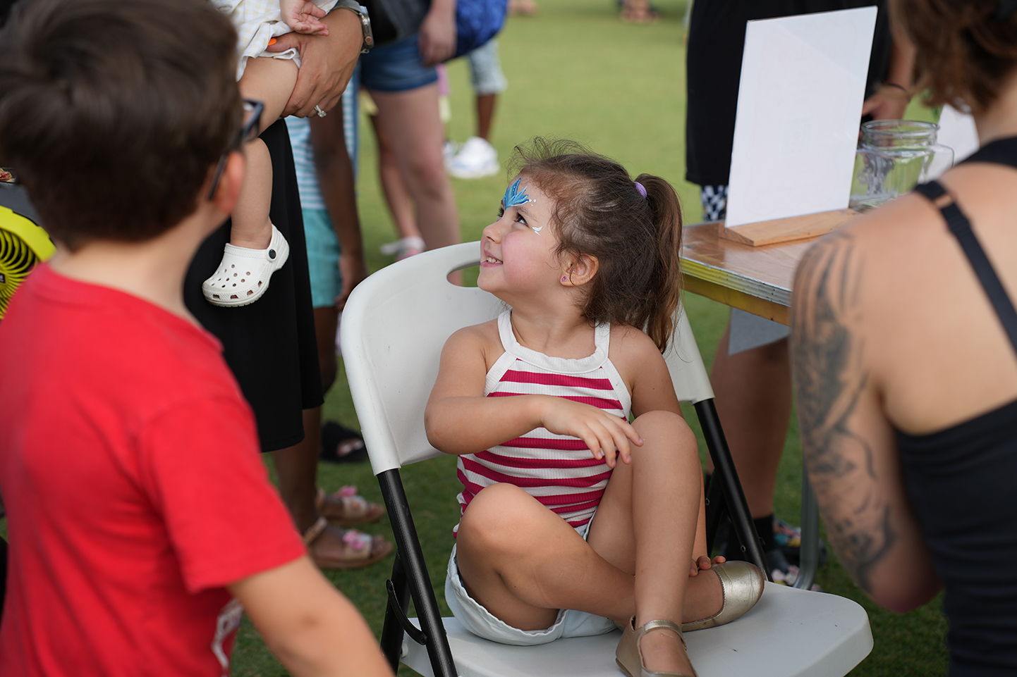 A young girl with blue face paint sits in a white chair, smiling, with a boy in red.
