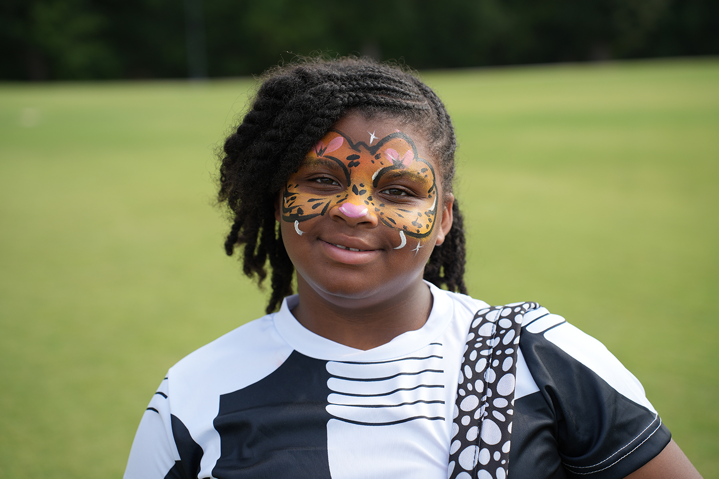 A girl with face paint showing a tiger's face stands on a grassy field.