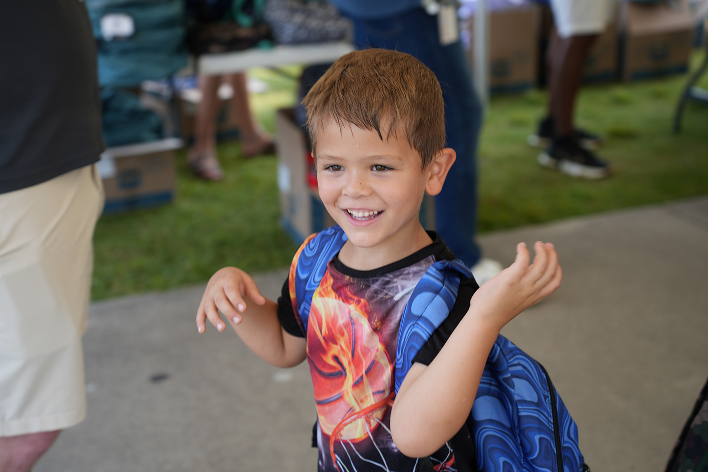 A smiling child with a backpack is standing outside near a table with various items.