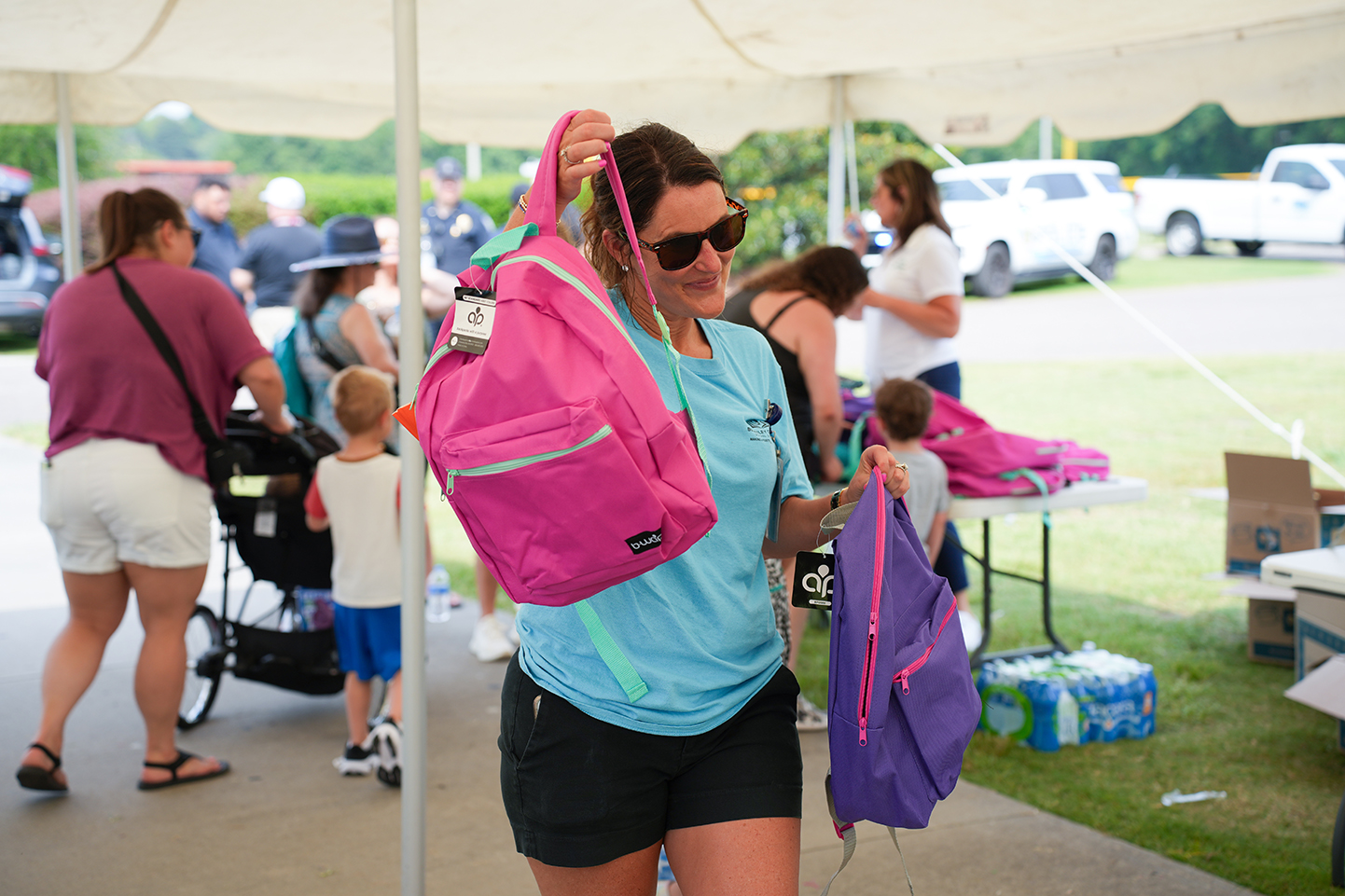 Woman in blue shirt and black shorts holds a pink backpack. Other people in the background.
