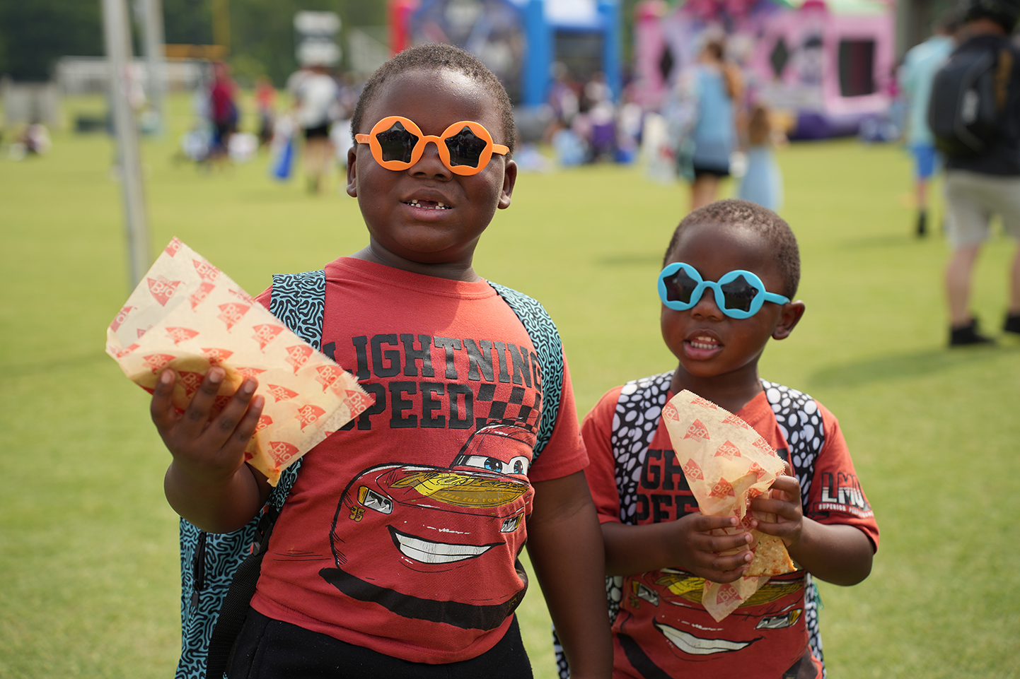 Two children stand on a field wearing matching glasses and holding food. A crowd of people, including adults, and inflatable structures in the background.