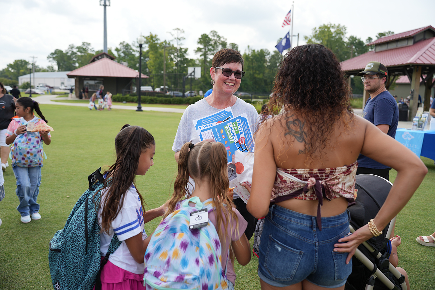 Adults and children gather in a park. One woman stands with a backpack and holds a book.