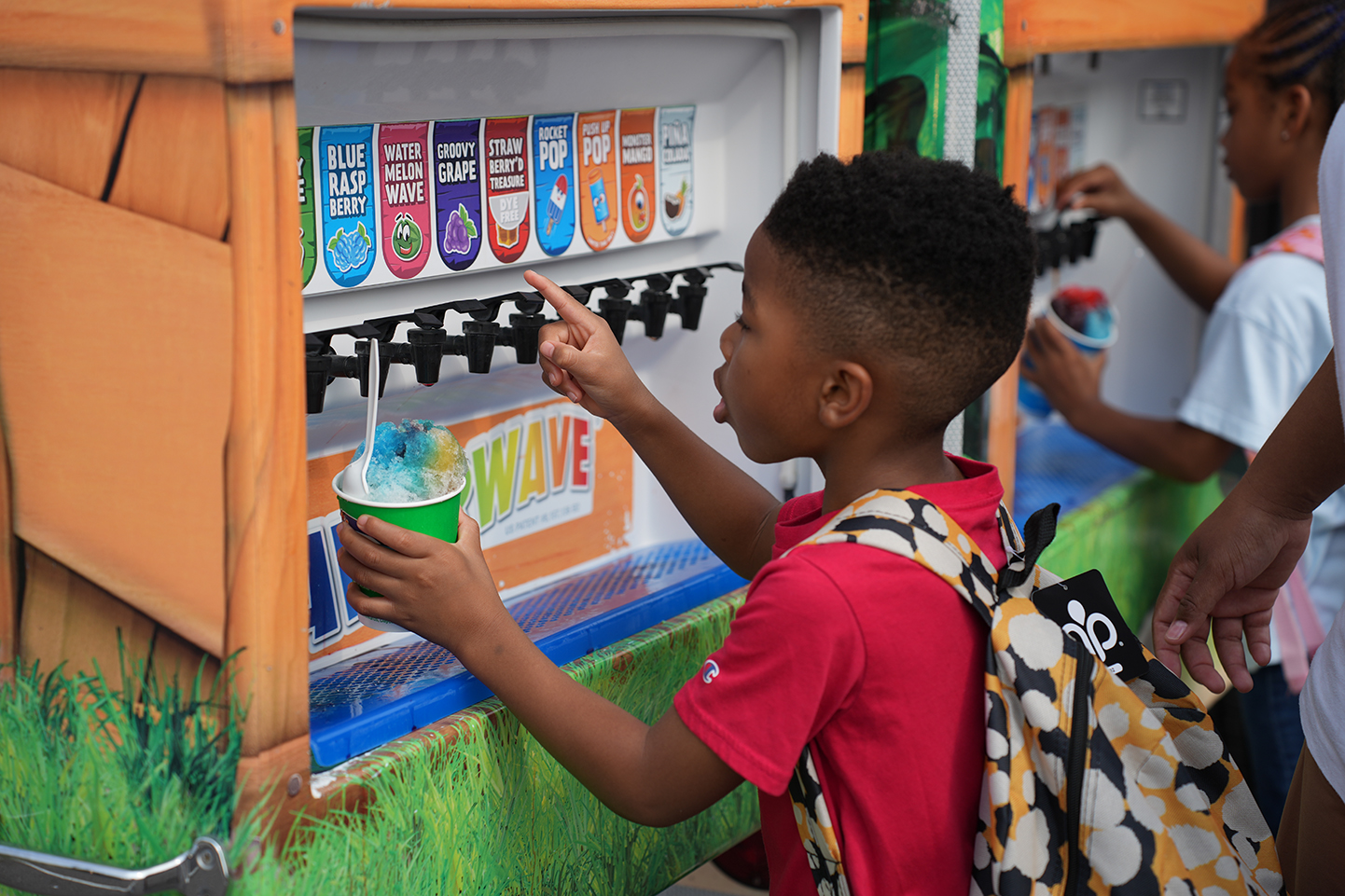 A young boy in a red shirt scoops ice cream from a dispenser. A backpack is on his back.