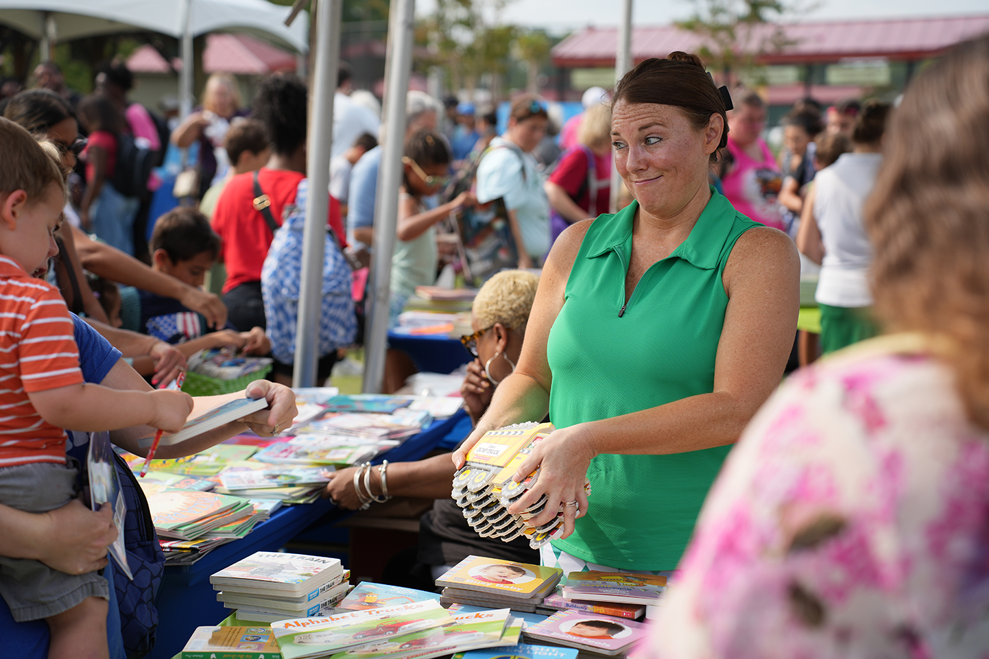 A woman in a green sleeveless shirt stands at a table, surrounded by people, some holding books.
