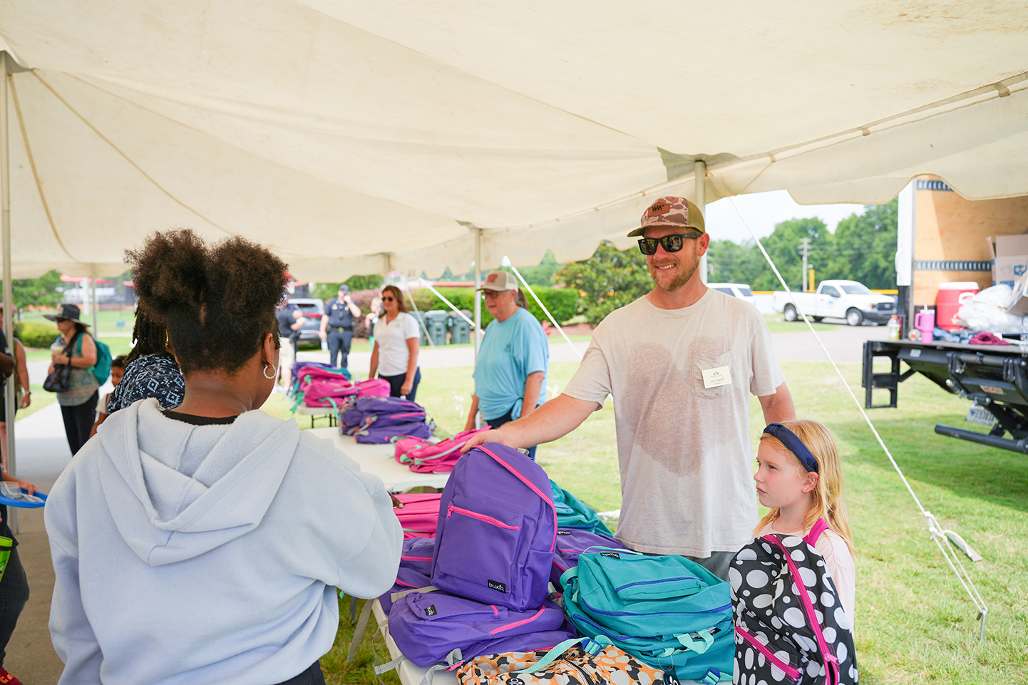 People are inside a tent under a white canopy. A man is giving a young girl a backpack.