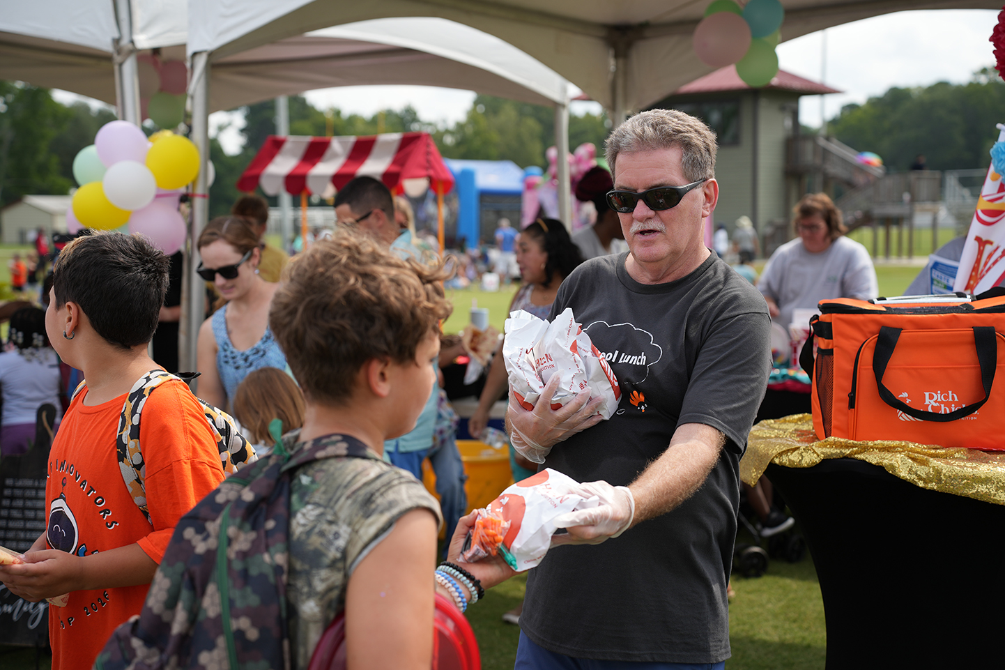 People at a community event with a man holding food. Tents, balloons, and a table with an orange bag are in the background.