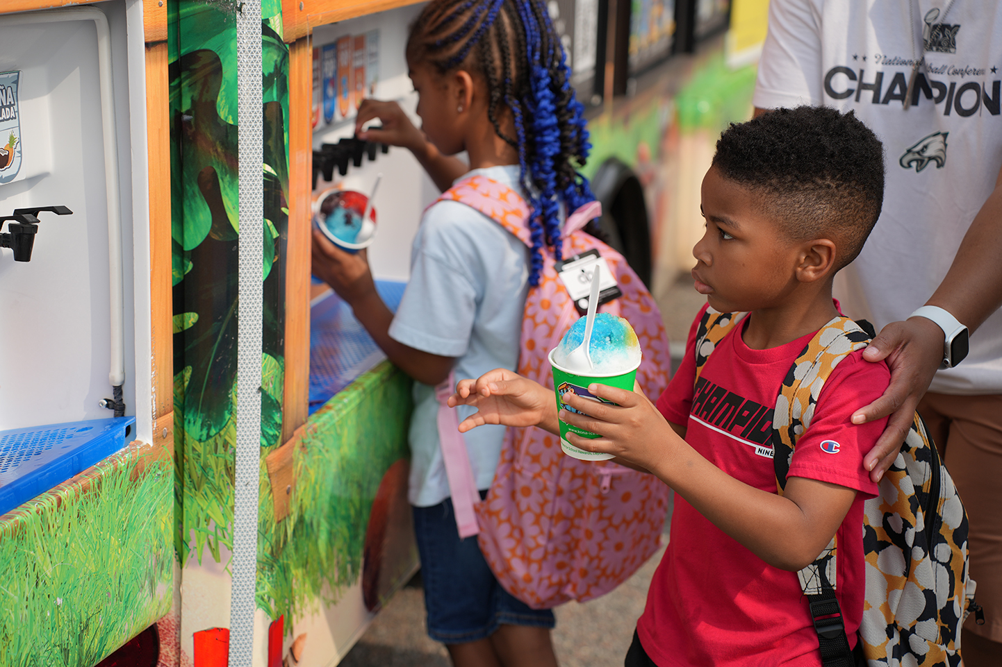 Two children with backpacks at an ice cream truck. One has a cup of ice cream.