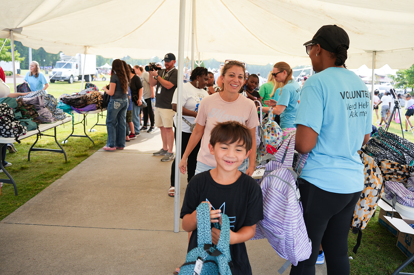 People under a tent. A smiling boy holds a backpack. An adult with bags stands nearby.