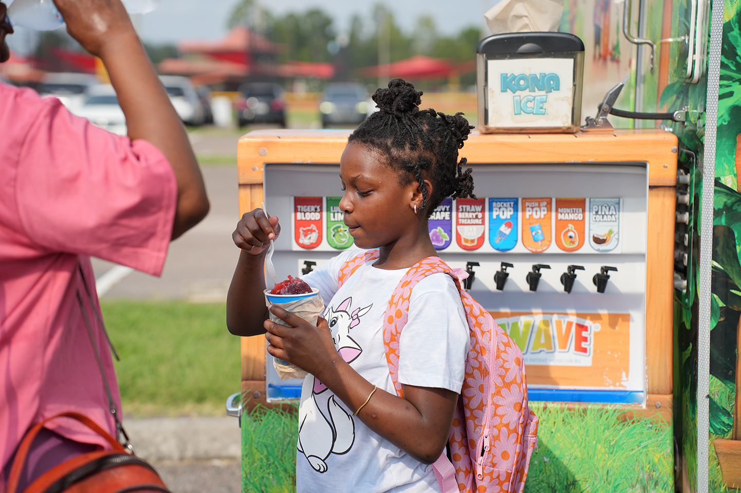 A young girl with a backpack eats ice cream from a machine outside. Another person stands behind her.