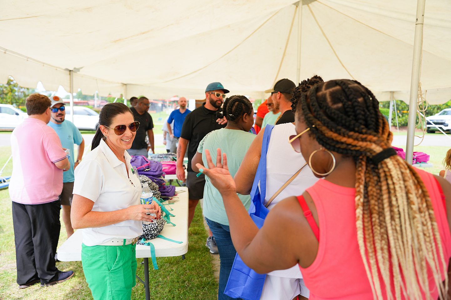 People under a white tent; a woman in sunglasses talks to a woman with braided hair.