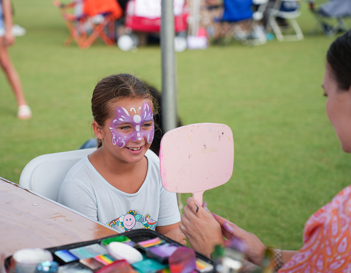 A young girl with a purple butterfly face paint sitting at a table while a woman applies face paint.