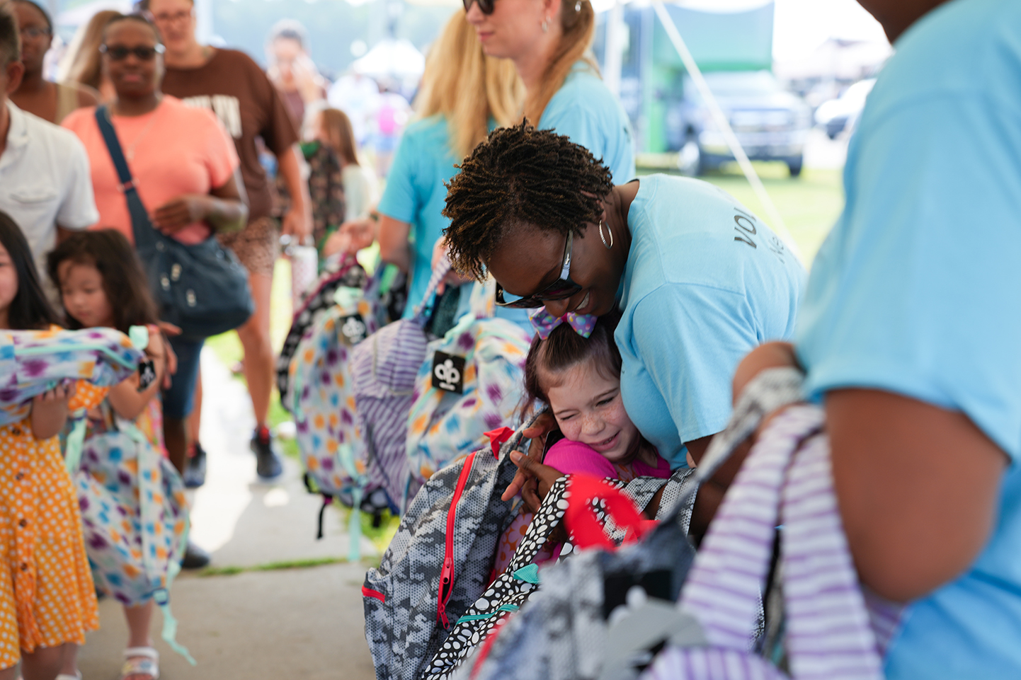 A woman with a backpack helps a young girl wearing a pink dress, both in a group of people.