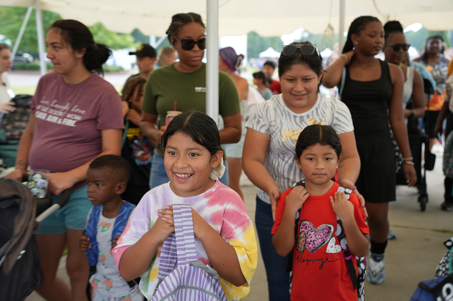 A group of children and adults walk through an outdoor area; two young girls in the front smile.