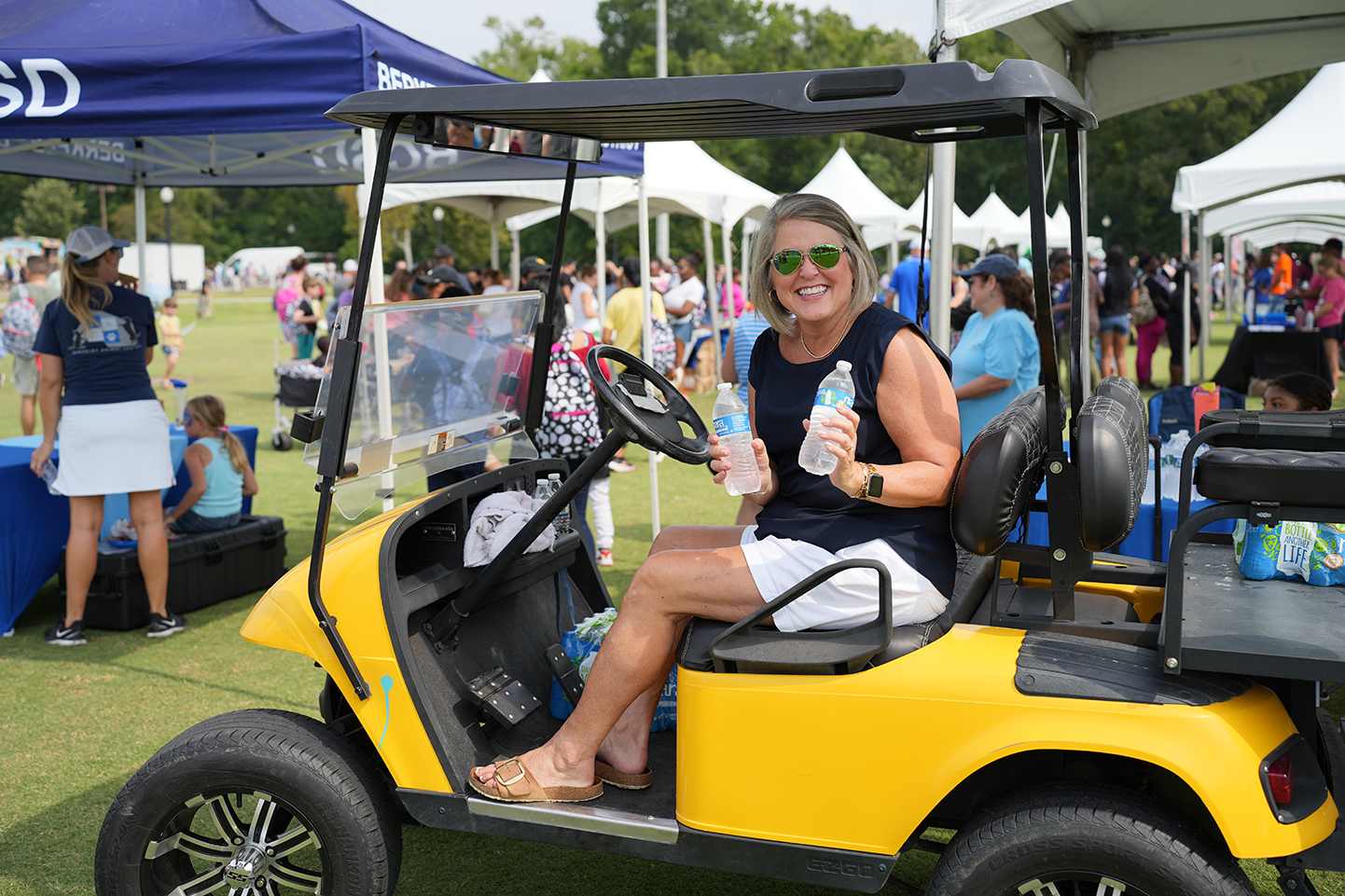 Woman in a yellow golf cart holding water bottles, with a blue tent and white canopy in background.