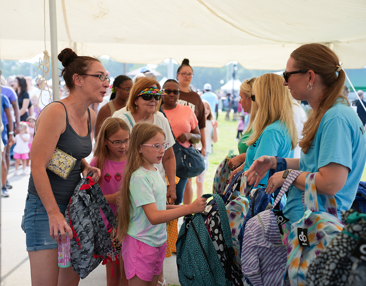 Several women and children gather under a white tent. Women hold backpacks and converse. Children examine bags. Many people are in the background.
