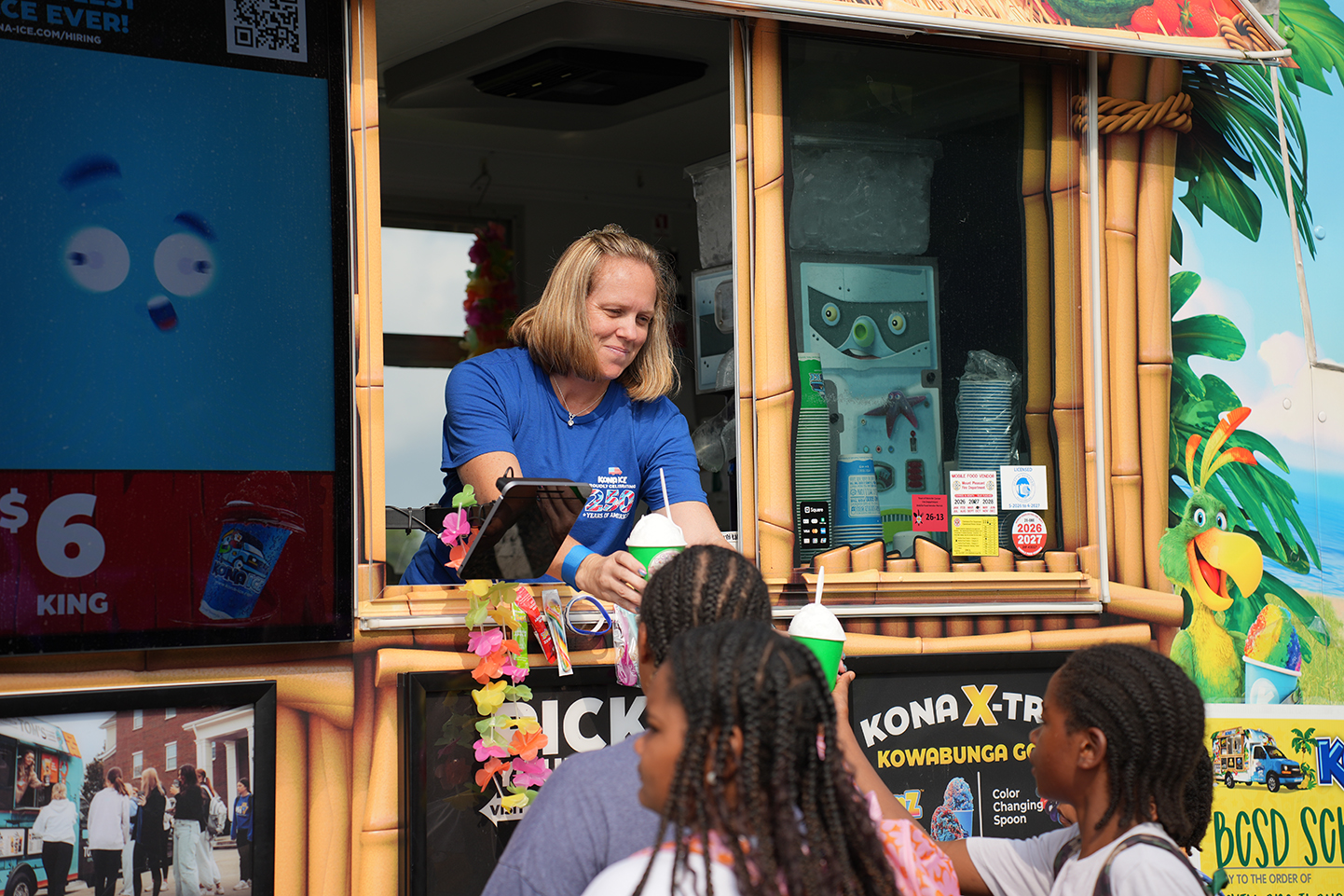 A woman stands inside a decorated food truck, smiling, while four children stand in front of her.