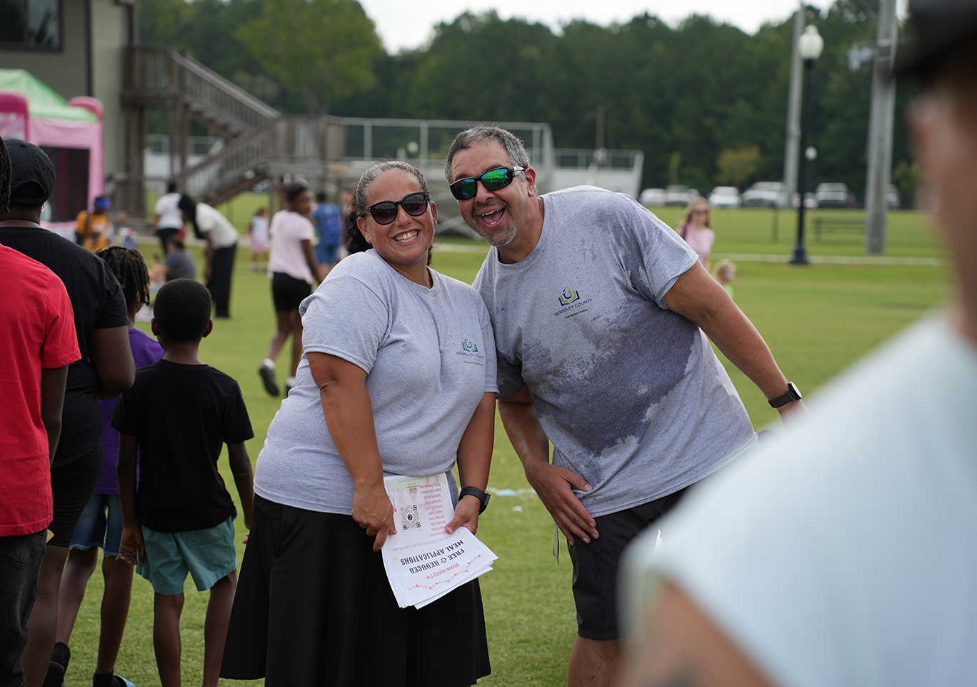 A smiling couple stands together on a grass field, with a group of people in the background.