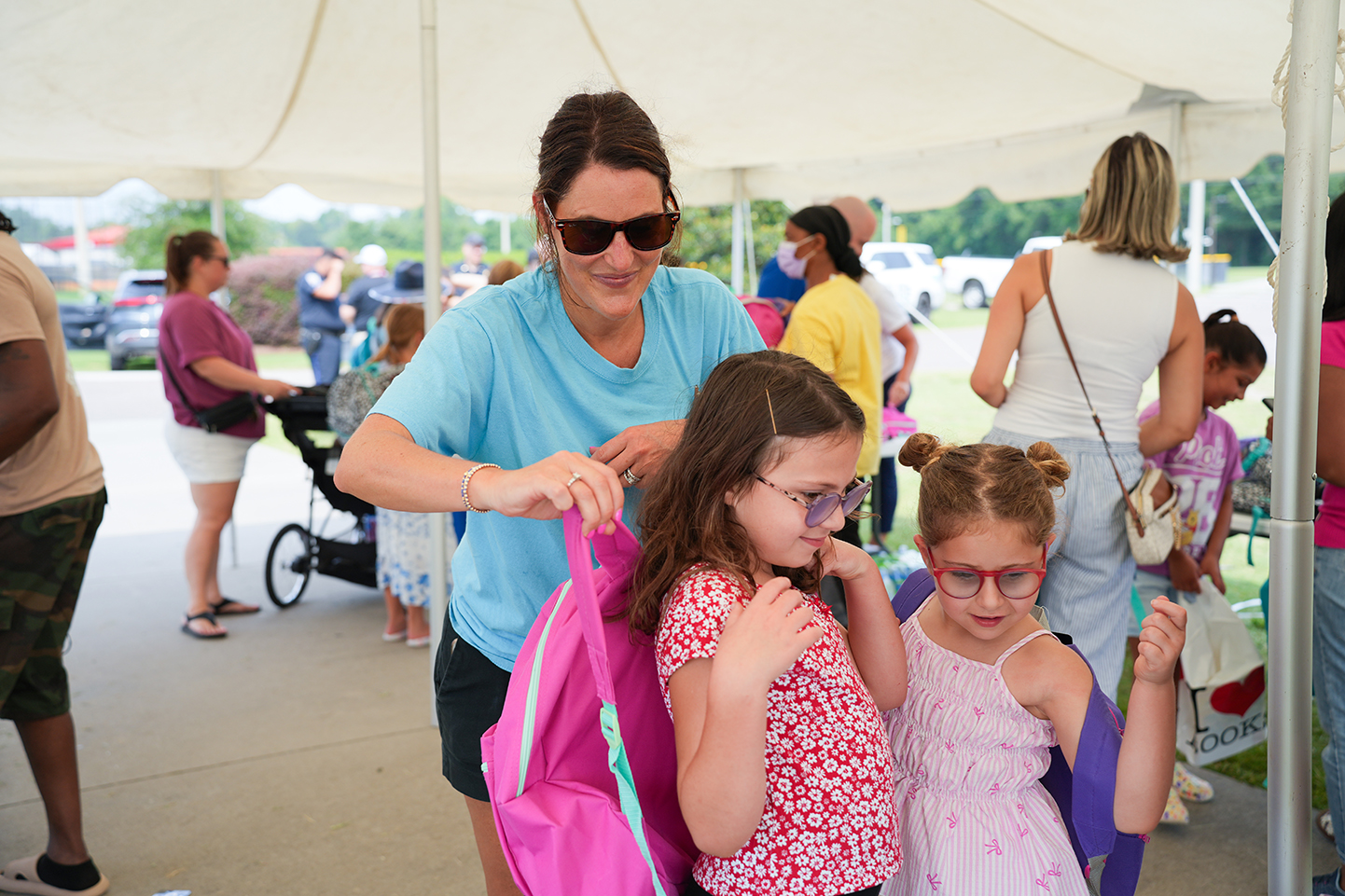 A woman helps two young girls with their backpacks at an outdoor event. The girls wear glasses.