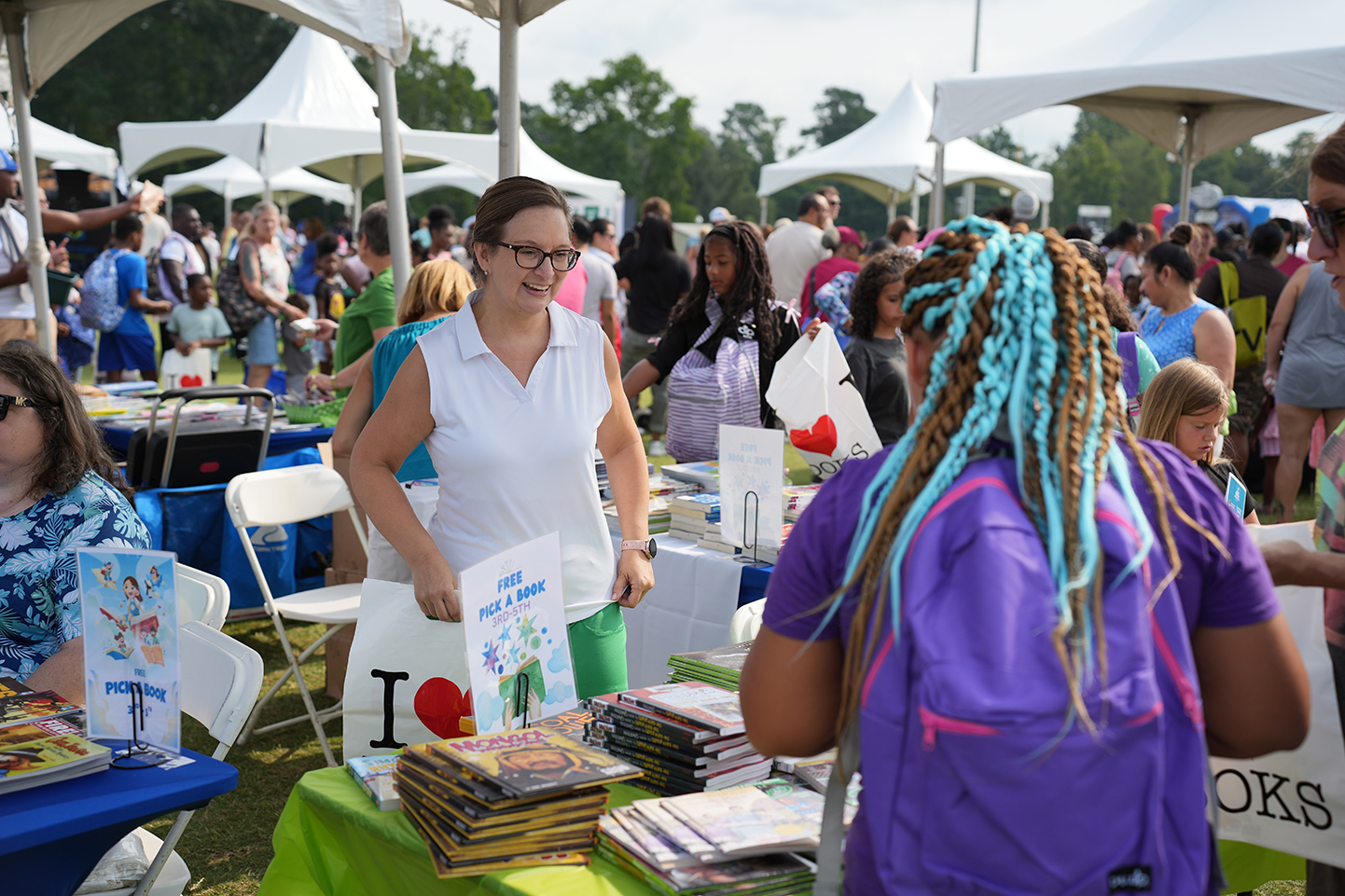 A crowd of people in an outdoor area with white tents. A woman in a white shirt stands by a table with books.