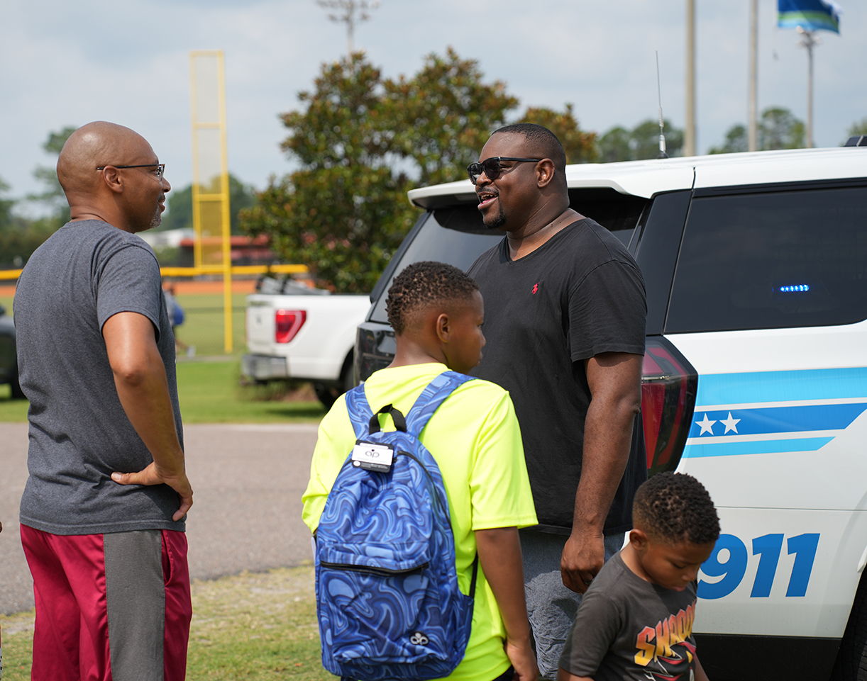 Three people near a white police vehicle; two adults and a child with a backpack. The vehicle has the number 911.