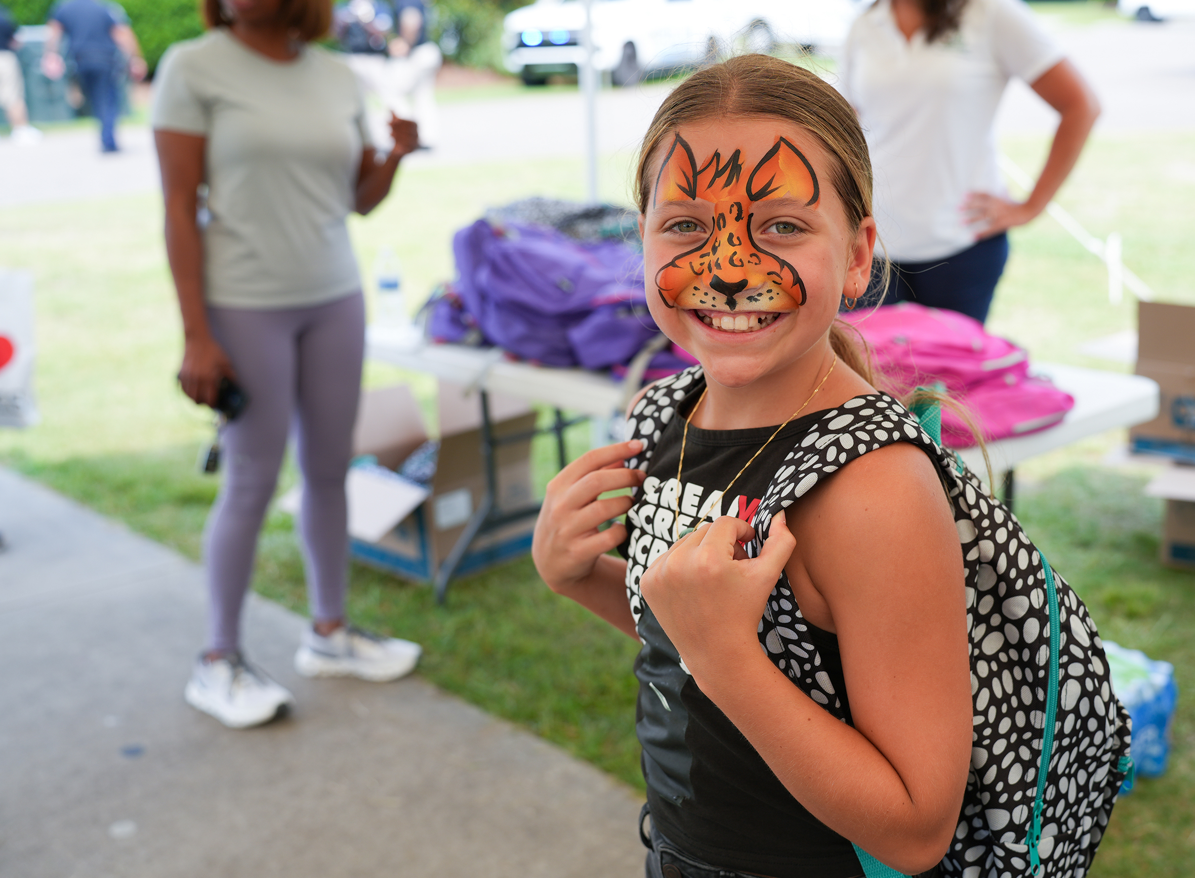 A girl with a lion face paint on smiles at the camera, holding a backpack. Two women stand behind.