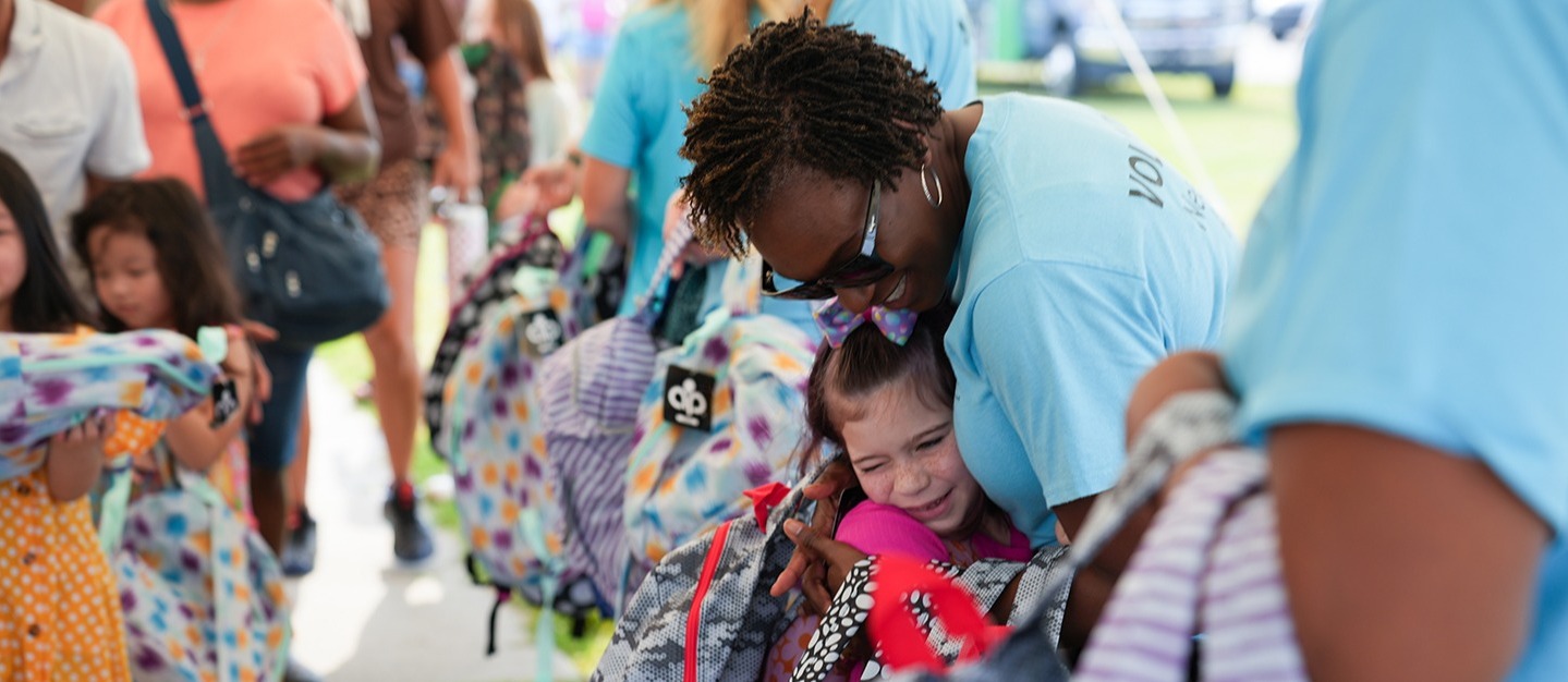 Adults and children gather, some wearing light blue shirts. A woman helps a child with a backpack.