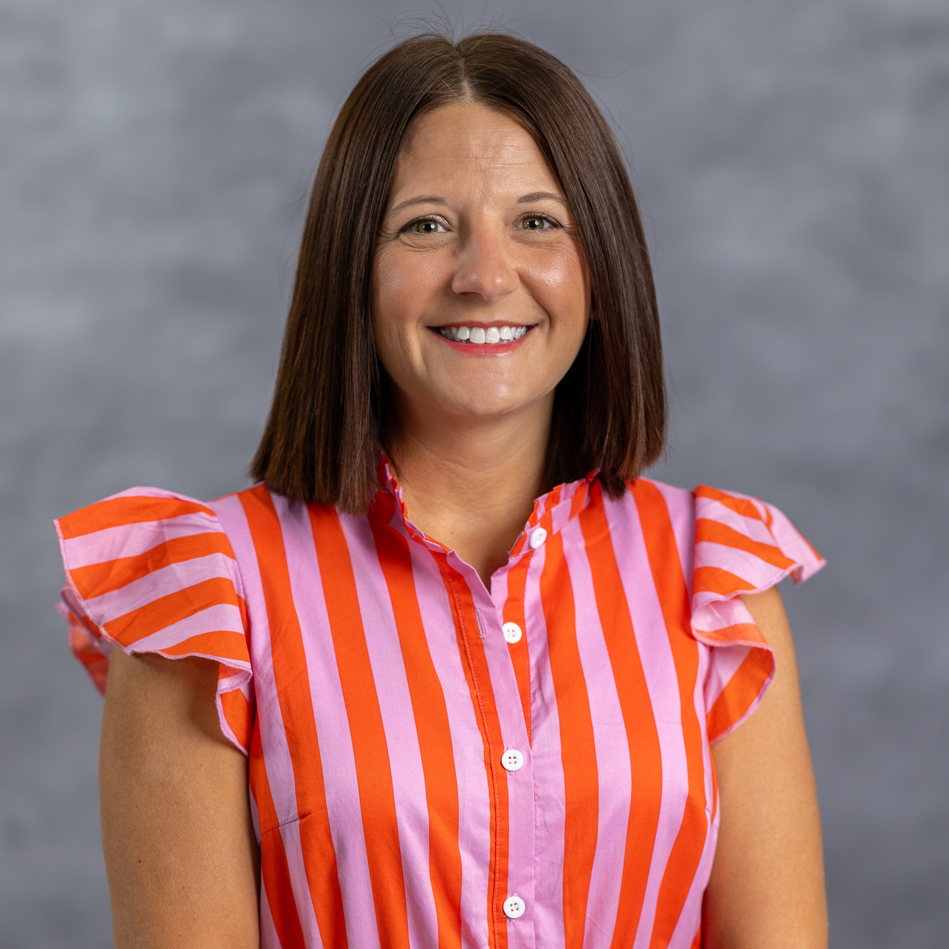 Woman smiling with short brown hair in a pink and orange striped shirt with ruffled sleeves.