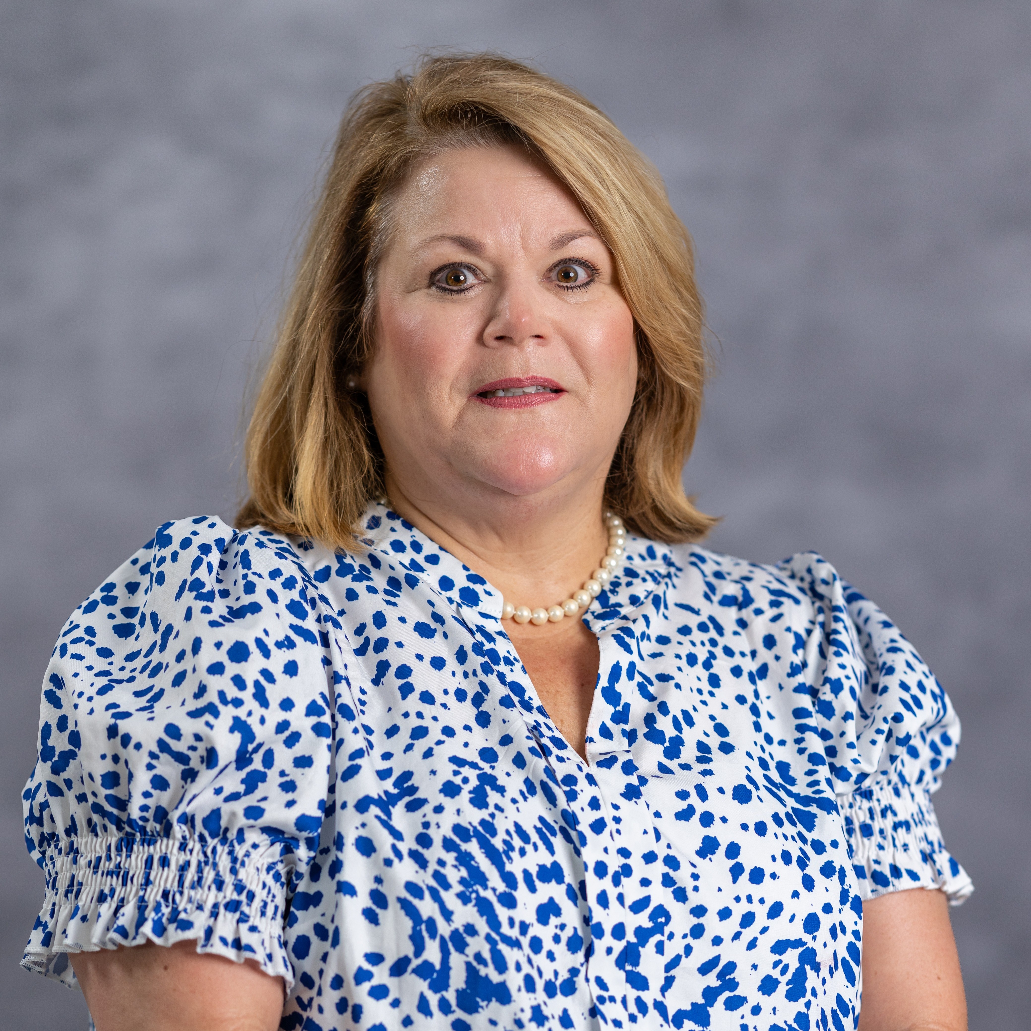 A woman with short blonde hair wearing a blue-white patterned blouse and a pearl necklace stands in front of a gray background.