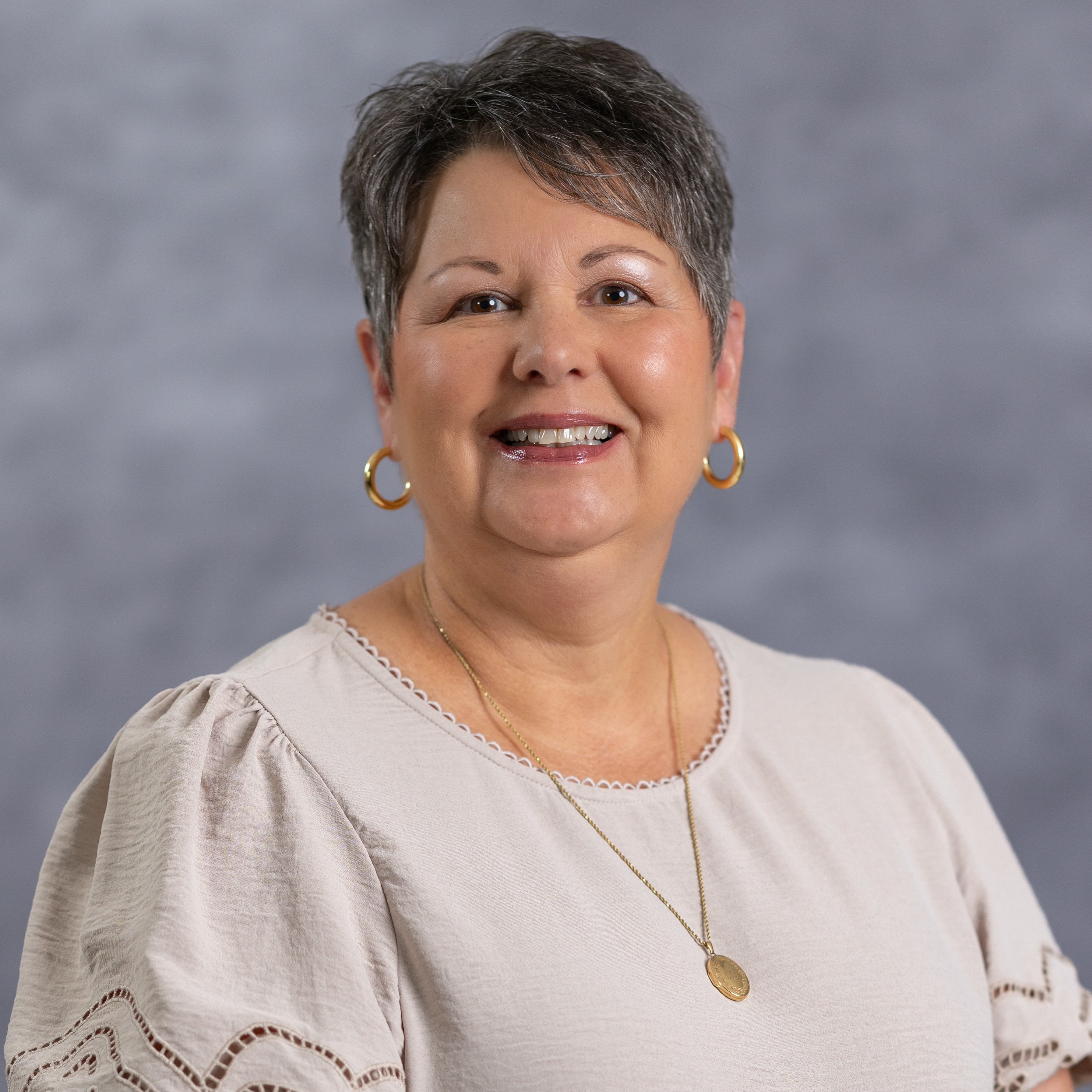 A woman with short hair and a smile poses for a photo. She wears a cream blouse with decorative lace and a gold pendant necklace.