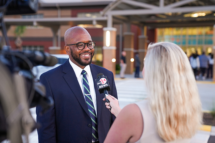 A man in a suit interviews a woman holding a microphone, with microphones and cameras behind them.