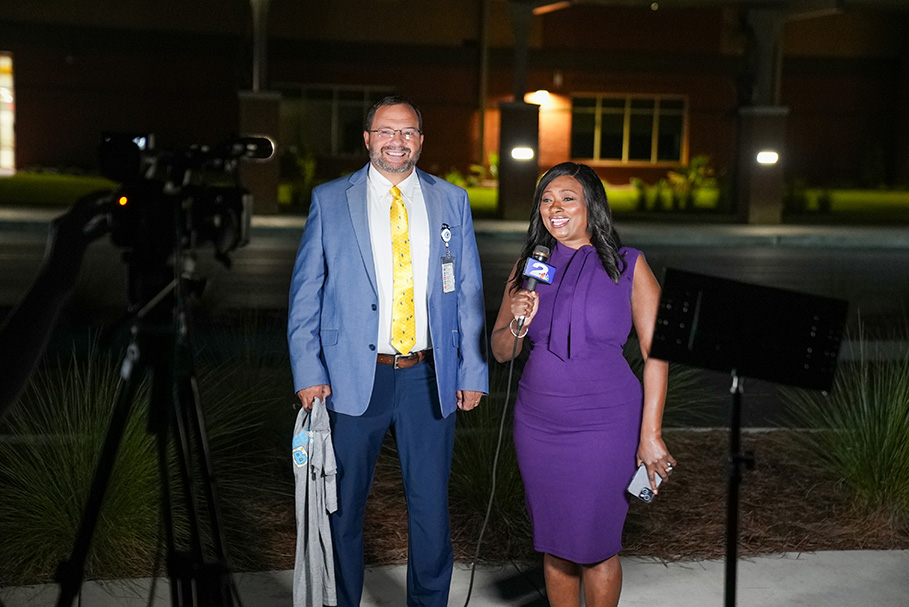 A man in a blue suit and yellow tie and a woman in a purple dress hold a microphone.