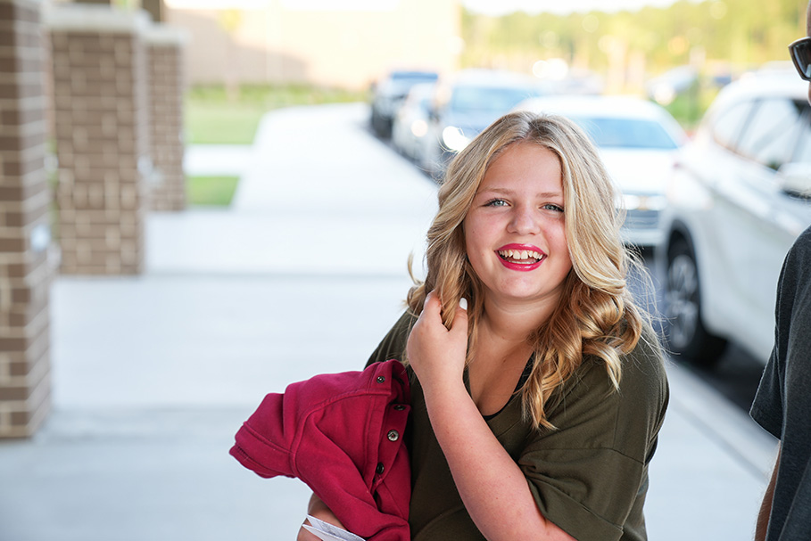 A girl smiles, holding a red jacket, on a sidewalk with parked cars in the background.