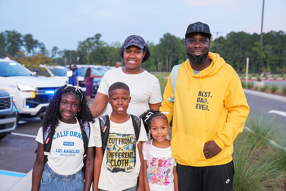 Family of four, consisting of two kids and two adults, stands in front of parked cars in a parking lot.