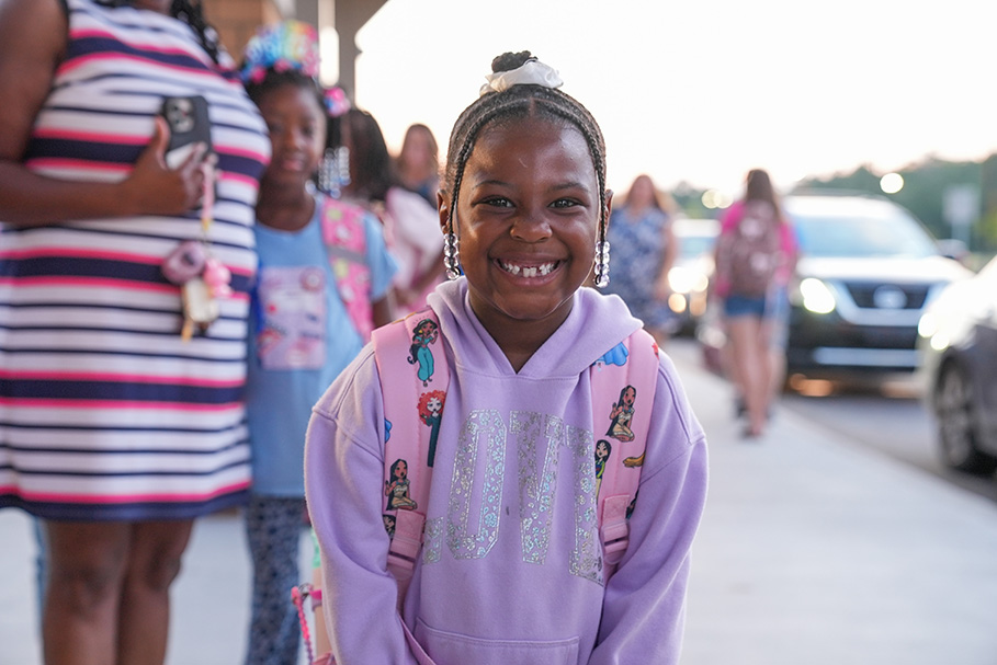 A girl in a purple hoodie with a backpack smiles at the camera, with other children and adults in the background.