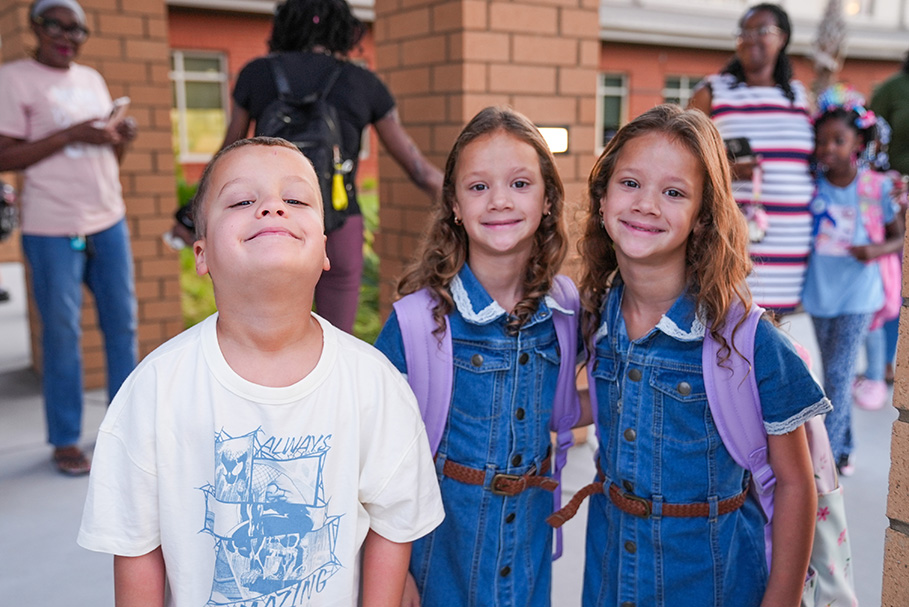Three children stand together, two girls in denim dresses and a boy in a white shirt. They smile.