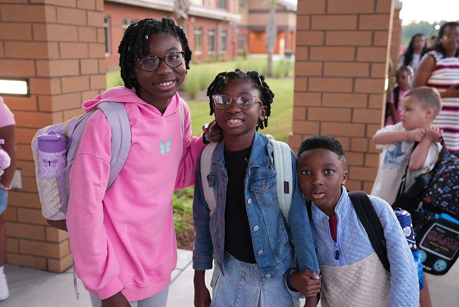 Three students, two girls and one boy, stand outside. A girl in a pink hoodie smiles.