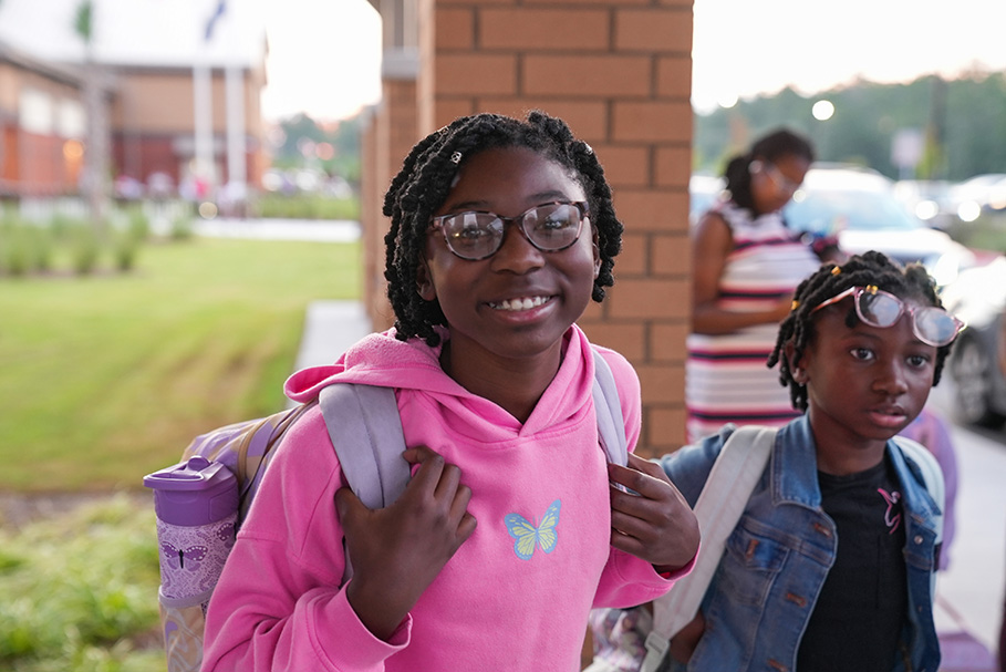 Two young girls stand outside. One wears a pink hoodie, glasses, and a backpack. The other wears a denim jacket and glasses. A building, grass, and parked cars are in the background.