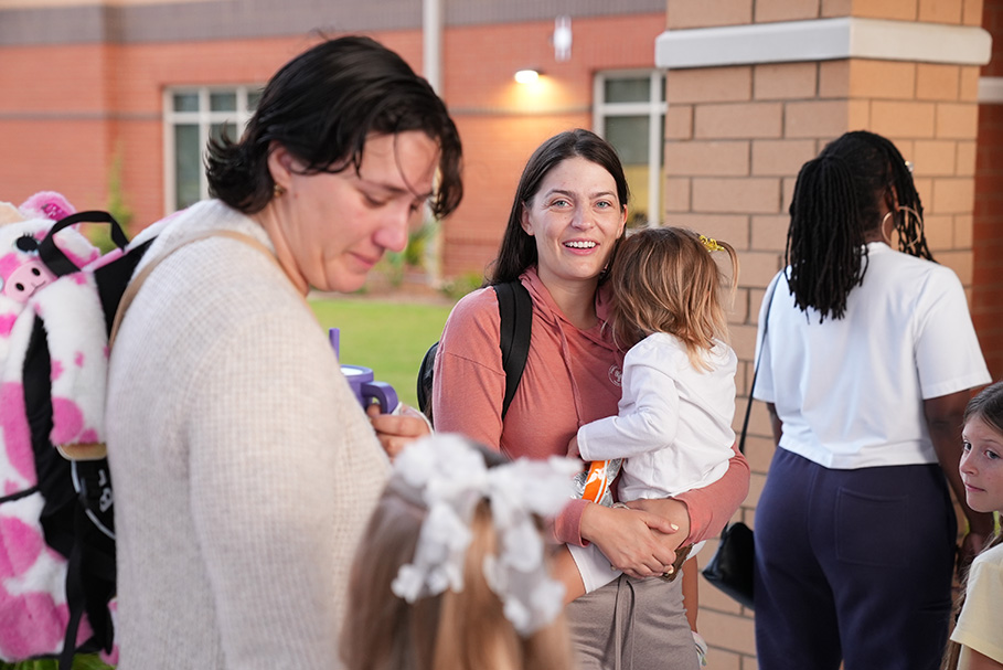 A group of adults and children in a hallway. Two adults look at a child with a pink backpack.