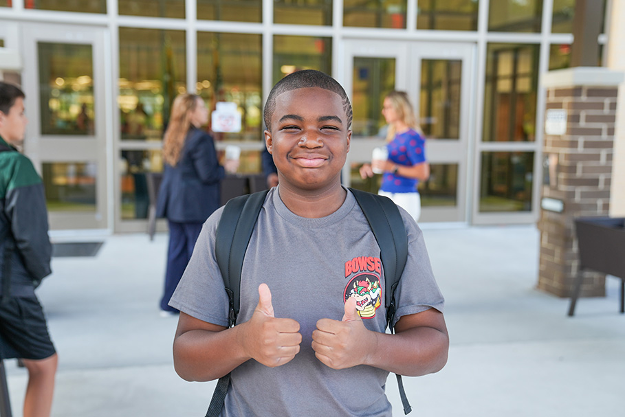 A boy in a gray T-shirt and a backpack gives two thumbs up in front of a school building.