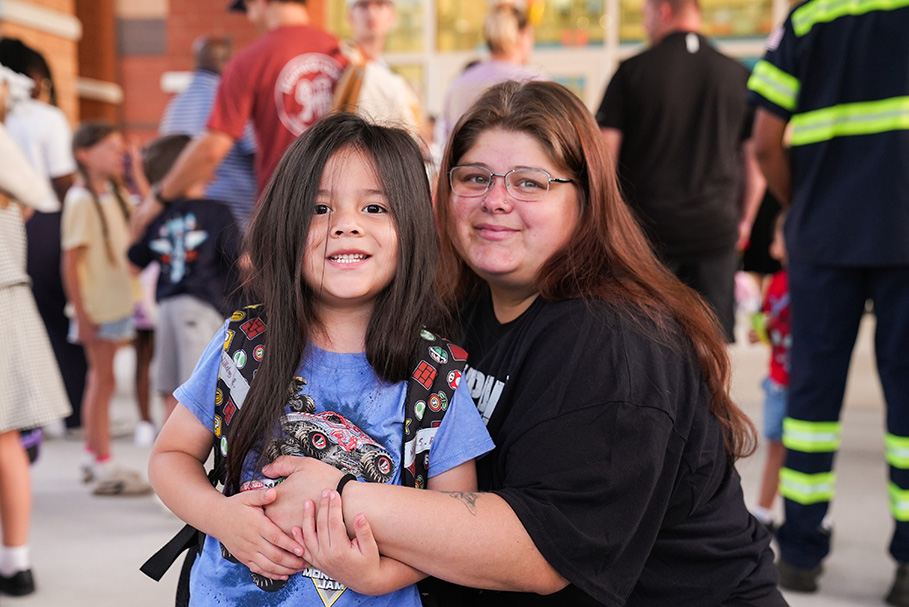 A woman hugs a smiling child with a backpack in a busy outdoor area with people.