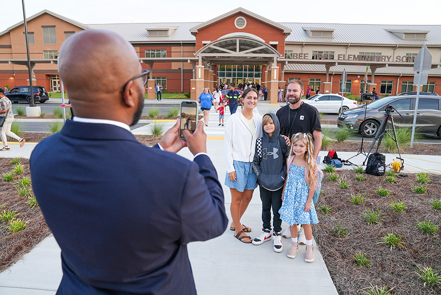 Man taking photo of family in front of a school building with a car parked behind.