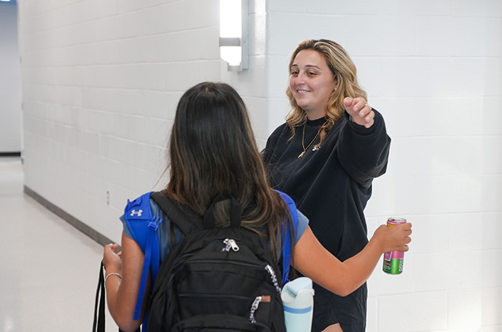 Two women in a hallway. One has a backpack, the other holds a can. Both are smiling.