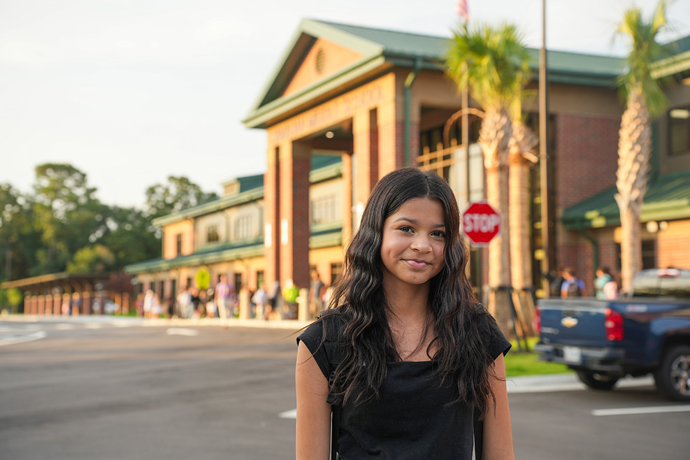 A young woman stands outside a building with palm trees, a blue pickup truck, and a stop sign.