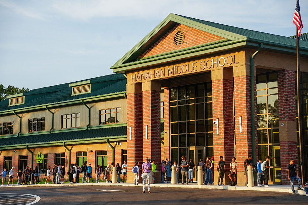 Exterior of a modern school building with large glass doors and a flagpole, with people standing in line outside.