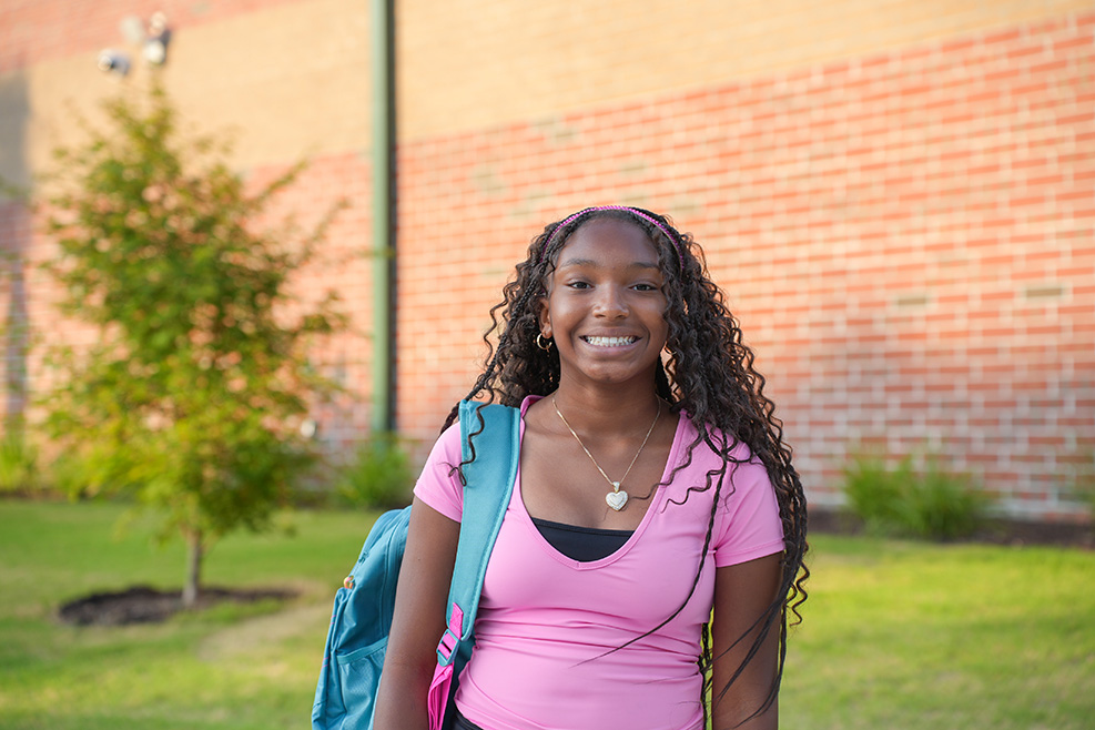 A girl with curly hair smiles in front of a red brick wall. She wears a pink shirt, earrings, and a blue backpack.