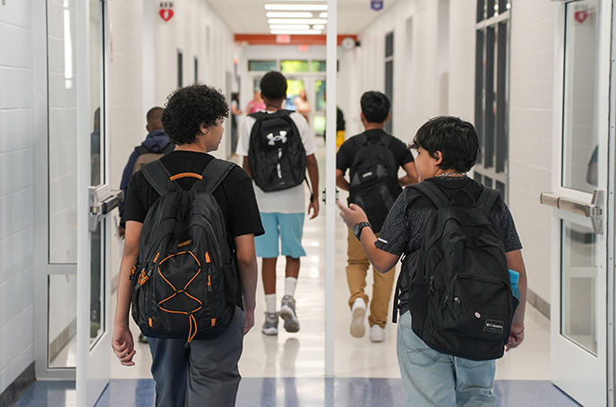 Several students with backpacks walk down a school hallway. Some are talking while others walk ahead.