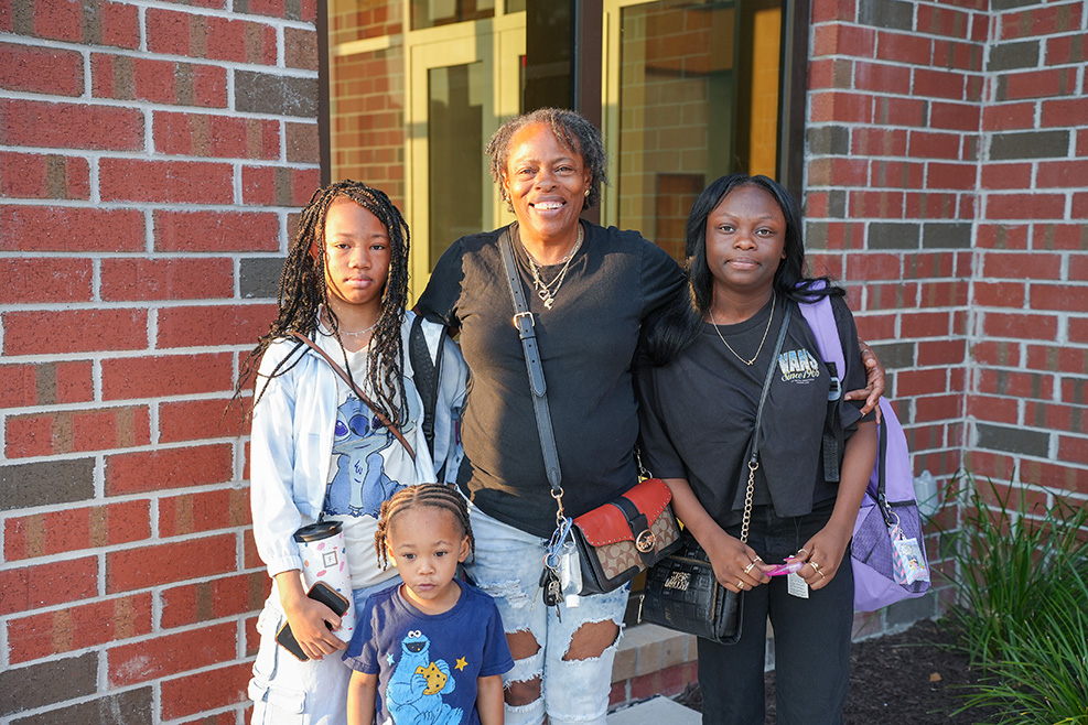 Four individuals standing outside a brick building, smiling and posing for a photo. Each holds a cup or bag.