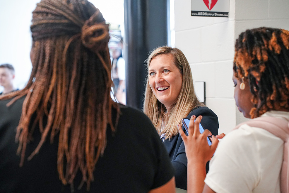 Three women talk in a room with white walls and a door. One woman smiles and holds a phone.