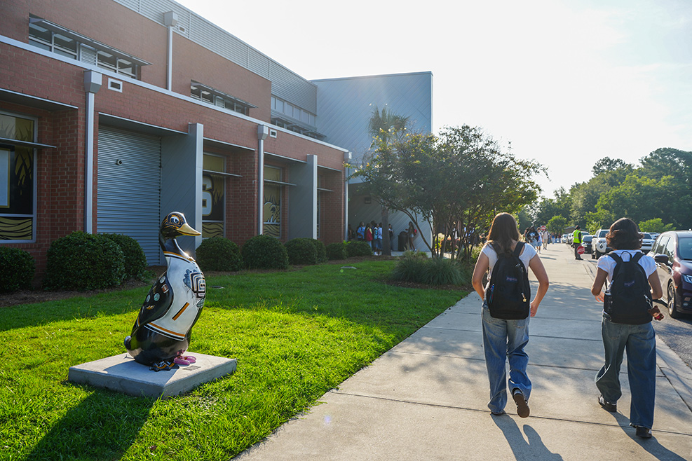 Two individuals with backpacks walk past a penguin statue in front of a building on a sunny day.